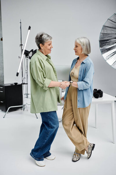 Two mature lesbian women in a photo studio, one posing as a model, the other taking photos.