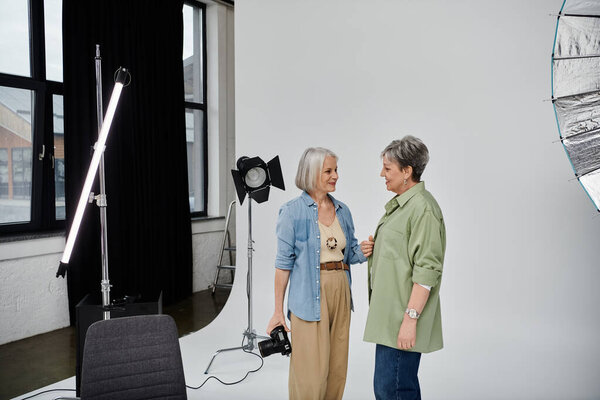 Two mature women in a studio setting, one holding a camera and the other posing.