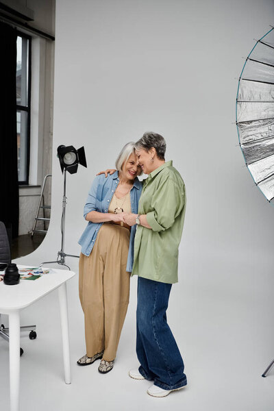 A mature lesbian couple poses for a photoshoot in a studio, embracing each other with a loving gaze.
