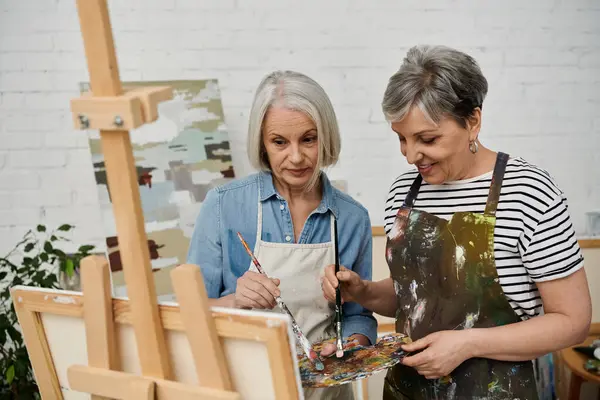 stock image Two mature women paint together in an art studio, one holding a palette and the other working on a canvas.