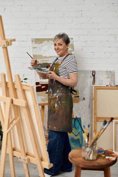 Женщина стоит в своей студии, кисть в руке, фокусируясь на палитре.