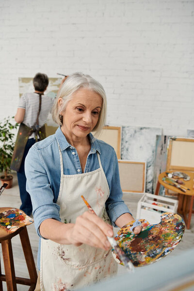 Пожилая женщина рисует в своей художественной студии, окруженная другими полотнами и художественными принадлежностями..