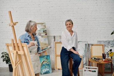 Gündelik giysiler içinde iki kadın, parlak bir sanat stüdyosunda resim seansına katılıyor..