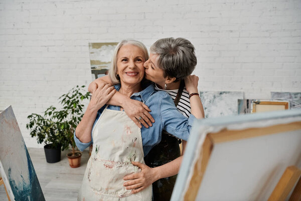 A lesbian couple, both mature women, embrace in an art studio, their love and connection palpable.