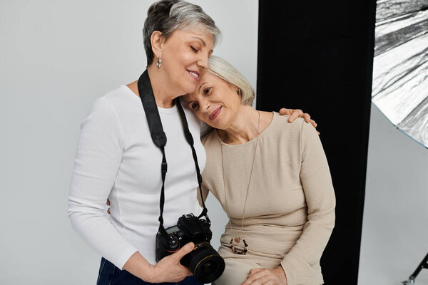 Two mature lesbians pose lovingly in a photo studio, one holding a camera, the other resting her head on her partner shoulder