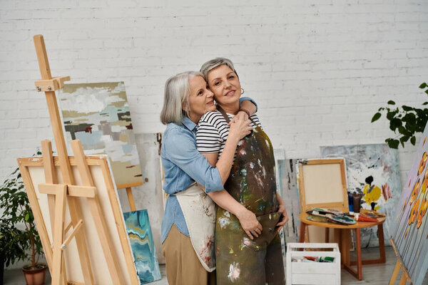 A lesbian couple embraces in an art studio, surrounded by canvases and paint.