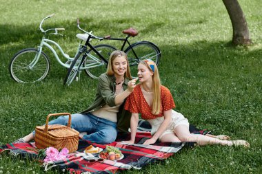 A young lesbian couple enjoys a picnic in a green park, feeding each other fruit and sharing a smile. clipart