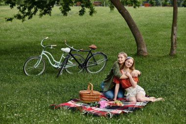 Genç bir lezbiyen çift yemyeşil bir parkta pikniğin tadını çıkarıyor, bisikletleri yakınlarda park halinde..