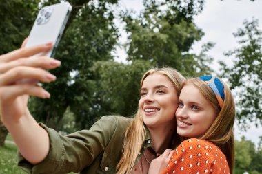 Parkta güneşli bir günün tadını çıkarırken iki genç kadın selfie çekmek için sarılıp gülümsüyor..