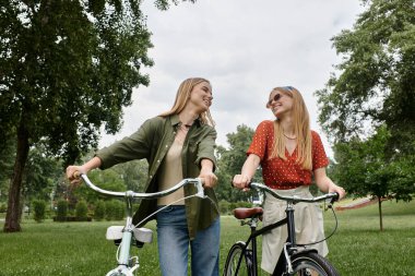 Genç bir lezbiyen çift yeşil bir parkta güneşli bir öğleden sonra bisiklet sürmekten zevk alıyor..
