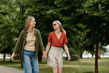 İki kadın yemyeşil bir parkta el ele yürür. Güneşli bir günün tadını çıkarırken gülümser ve gülerler..