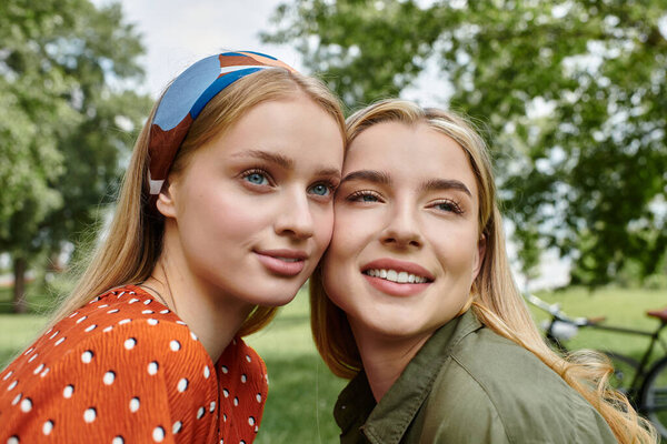Two young women, heads close together, smile as they share a moment of happiness in a sunny park.