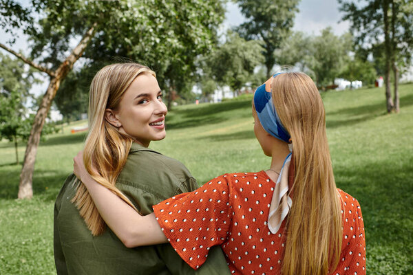 Two young women stroll hand-in-hand through a lush green park, their laughter echoing through the trees.