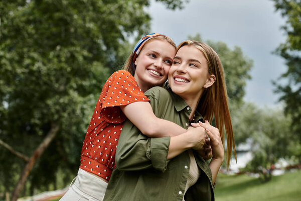 Two young women stand close together in a lush park, one wrapped in the arms of the other, both smiling brightly.