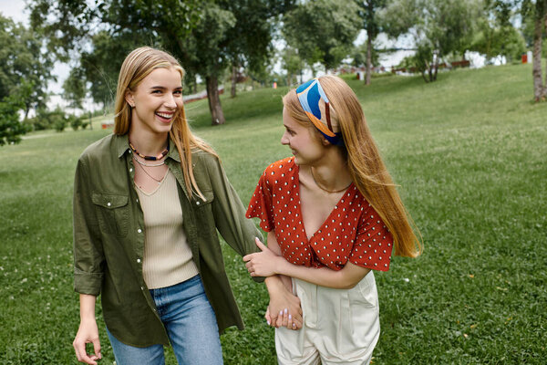 Two young women walk hand-in-hand through a lush green park, their laughter echoing through the trees.