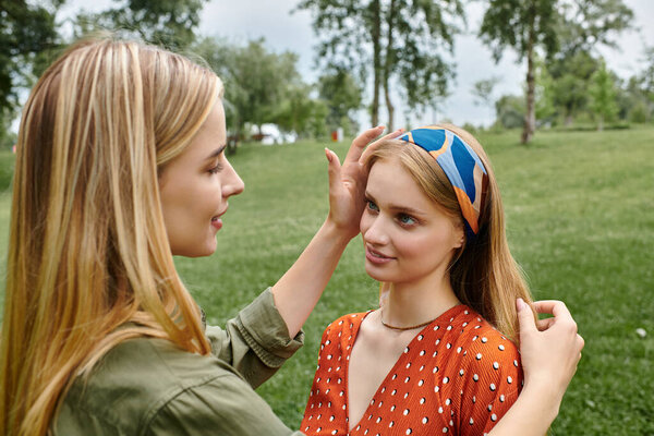 A young lesbian couple shares a tender moment in a green park, one woman gently stroking the other hair.