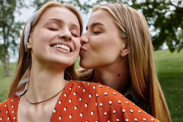 A young woman in a red polka dot dress smiles as her girlfriend kisses her cheek while on a date in a green park.
