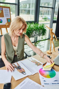 Yeşil yelekli ve beyaz bluzlu sarışın bir kadın masasında oturmuş moda çizimlerini ve renk örneklerini inceliyor..