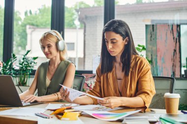 Profesyonel olarak giyinmiş iki kadın, modern ofis ortamında bir proje üzerinde çalışıyor, işbirlikçi ve odaklanmış bir ortam sergiliyorlar..