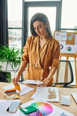 A woman in a mustard shirt examines a design sketch at a cluttered desk with color palettes, papers, and a smartphone clipart