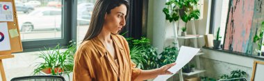 A woman in a beige blouse reviews papers, standing in a modern office with large windows and greenery. clipart