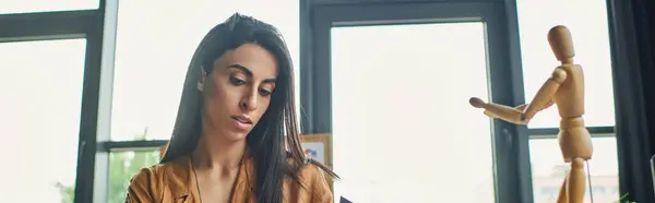 stock image A young woman with long dark hair looks thoughtfully at a wooden figure, lost in thought while working at her desk.