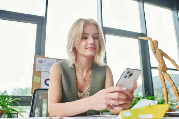 Молодая блондинка сидит за столом в светлом офисе и проверяет телефон..