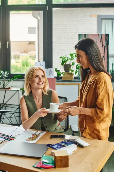 stock image Two women in a modern office, one offers a cup of coffee to the other while they work on a project together.