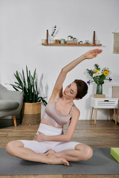 Азиатка в кукурузном топе сидит, скрестив ноги на коврике для йоги, тренируясь с протянутой над головой рукой..