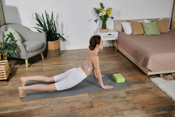 stock image A young woman practices yoga on a mat in a modern apartment, using a block for support.