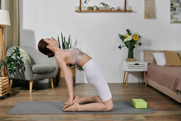 stock image A woman in a crop top practices yoga at home, kneeling on a mat with her head back, in a modern apartment.