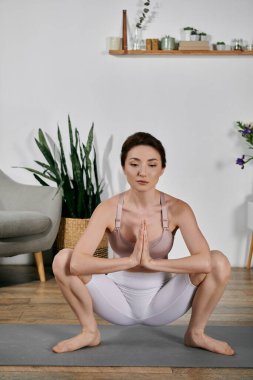 Ekin bluzlu ve şortlu bir kadın modern apartmanındaki yoga minderinde çömelmiş bir pozisyonda meditasyon yapıyor..