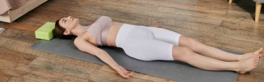 Uzun, koyu renk saçlı genç bir kadın yoga minderinde yatarken meditasyon yapıyor..