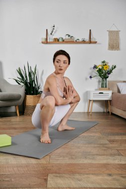 Ekin üstü giymiş Asyalı bir kadın modern bir dairede gri bir paspasın üzerinde yoga ve meditasyon yapıyor..