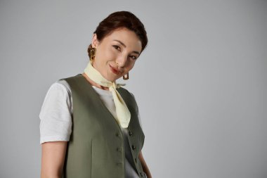 Yeşil yelek ve beyaz boyun atkısı takmış şık bir Asyalı kadın gri arka planda poz veriyor..