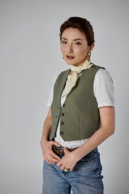Yeşil yelekli ve beyaz gömlekli şık bir Asyalı kadın gri bir fonda poz veriyor..