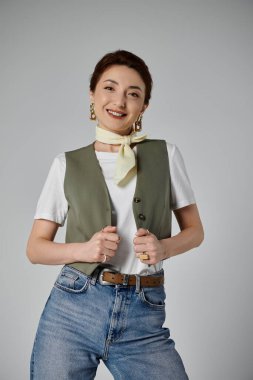 Yeşil yelekli, beyaz tişörtlü, sarı atkılı, kot pantolonlu, kahverengi kemerli, sıcak bir gülümsemesi olan Asyalı bir kadın.