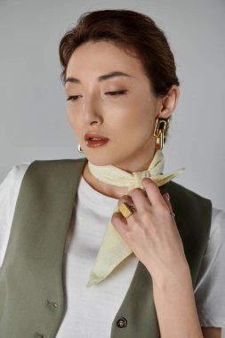 Beyaz tişört giyen şık bir Asyalı kadın ve sarı atkı gri arka planda poz veriyor..