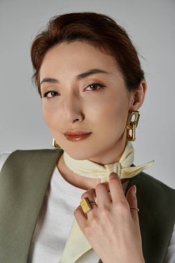 Yelek ve boyun atkısı takan güzel bir Asyalı kadın gri bir arkaplan karşısında kendine güvenen bir ifadeyle poz veriyor..