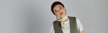 Yeşil yelek giyen şık bir Asyalı kadın gri bir fonda dikilirken kameraya poz veriyor..