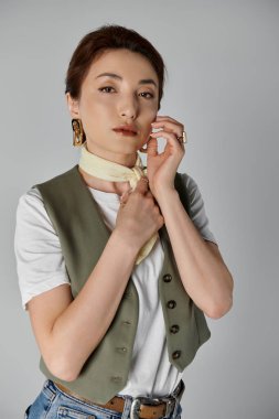 Asyalı kadın gri arka plana karşı poz veriyor, beyaz boyun eşarbını ayarlıyor. Yeşil yelek, onun günlük kıyafetine incelik katıyor.