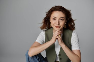 Şık bir Asyalı kadın gri bir arkaplana karşı poz veriyor, atkı ve yelek takıyor. Elleri önünde birbirine kenetlenmiş..