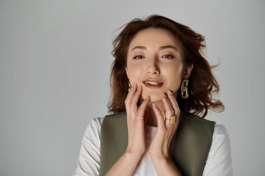 Yelek giymiş şık bir Asyalı kadın gri bir fonda poz veriyor, elleri nazikçe yüzüne dokunuyor..