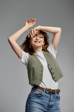 Şık giyinmiş, yelek ve kot pantolon giyen Asyalı bir kadın gri bir zemine poz veriyor. Gülüşü neşe ve güven saçıyor..