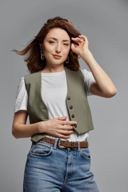 Yeşil yelekli şık Asyalı kadın, beyaz tişört, mavi kot pantolon, kahverengi kemer, gri arka planda poz, saç akışı, yüze yakın el.
