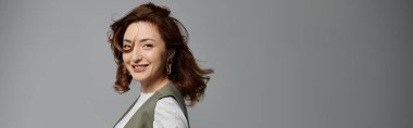 Siyah saçlı, şık bir yelek giyen, gri bir arka plan önünde poz veren, saçları akan ve gülüşü parlak bir kadın..