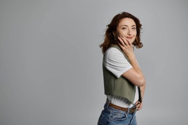 Kahverengi saçlı, şık bir Asyalı kadın gülümseyip kameraya poz veriyor. Yeşil yelek, beyaz gömlek ve kot pantolon giyiyor..