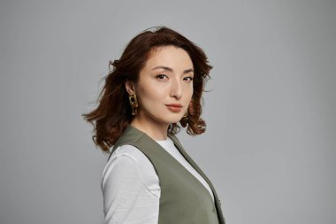 Koyu kahverengi saçlı güzel bir Asyalı kadın gri arka planda yeşil bir yelek giyiyor..