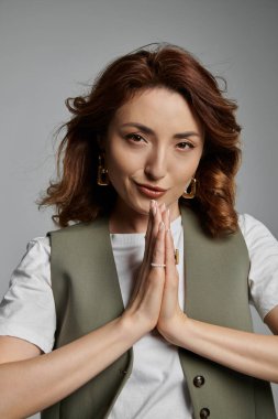 A stylish Asian woman in a green vest with a soft smile stands against a gray background, her hands clasped in prayer. clipart