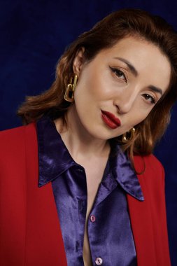 Asyalı bir kadın şık kırmızı bir takım elbise ve açık ipek bir gömlek giyer, koyu mavi bir arkaplanda poz verir..